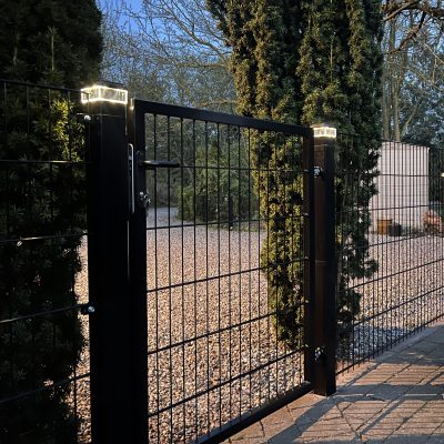 Et metallåge 2D panelfelt 6/5/6 mm sort Ral 9005 komplet med solcellelamper oven på stolperne, placeret blandt høje stedsegrønne træer og en grusjord, fotograferet i skumringen.