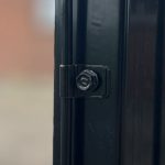 Nærbillede af en sort metaloverflade, der viser en sekskantet bolt og låsesamling sikkert fastgjort med Panelclips - Panel/Låge Fastgørelsesclips. Baggrunden er ude af fokus og viser en dæmpet, utydelig farve, der ser ud til at være en væg eller anden overflade.