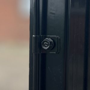 Nærbillede af en sort metaloverflade, der viser en sekskantet bolt og låsesamling sikkert fastgjort med Panelclips - Panel/Låge Fastgørelsesclips. Baggrunden er ude af fokus og viser en dæmpet, utydelig farve, der ser ud til at være en væg eller anden overflade.