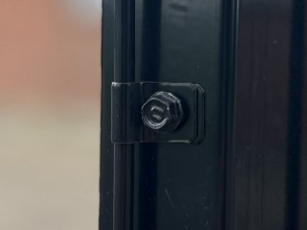 Nærbillede af en sort metaloverflade, der viser en sekskantet bolt og låsesamling sikkert fastgjort med Panelclips - Panel/Låge Fastgørelsesclips. Baggrunden er ude af fokus og viser en dæmpet, utydelig farve, der ser ud til at være en væg eller anden overflade.