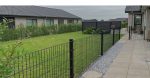 En baggård med en velholdt græsplæne, omkranset af et trådhegn og en række buske. Terrassen, belagt med betonfliser, føles hyggelig under den overskyede himmel. To huse med mørke tage er synlige, mens en lille grussti løber langs panelhegnen.