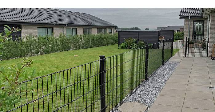 En baggård med en velholdt græsplæne, omkranset af et trådhegn og en række buske. Terrassen, belagt med betonfliser, føles hyggelig under den overskyede himmel. To huse med mørke tage er synlige, mens en lille grussti løber langs panelhegnen.
