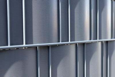 Et nærbillede af en Renovationsskjuler EcoHide Komplet med vertikalt ribbede metalpaneler. Panelerne er justeret i et gittermønster, hvilket skaber rektangulære sektioner mellem vandrette og lodrette stænger. Overfladen har en let reflekterende kvalitet, der kaster svage skygger.