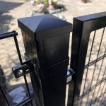 Nærbillede af en Metallåge 2D Panelfyld 6/5/6mm Sort Ral 9005 Komplet hegnspæl med en firkantet kappe og trådhegn fastgjort, placeret udendørs på stenbelægning med blødt slørede planter i baggrunden.