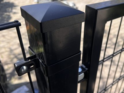 Nærbillede af en Metallåge 2D Panelfyld 6/5/6mm Sort Ral 9005 Komplet hegnspæl med en firkantet kappe og trådhegn fastgjort, placeret udendørs på stenbelægning med blødt slørede planter i baggrunden.
