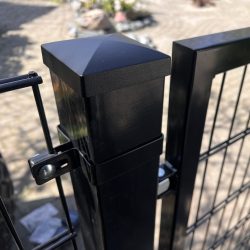 Nærbillede af en Metallåge 2D Panelfyld 6/5/6mm Sort Ral 9005 Komplet hegnspæl med en firkantet kappe og trådhegn fastgjort, placeret udendørs på stenbelægning med blødt slørede planter i baggrunden.