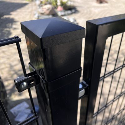 Nærbillede af en Metallåge 2D Panelfyld 6/5/6mm Sort Ral 9005 Komplet hegnspæl med en firkantet kappe og trådhegn fastgjort, placeret udendørs på stenbelægning med blødt slørede planter i baggrunden.