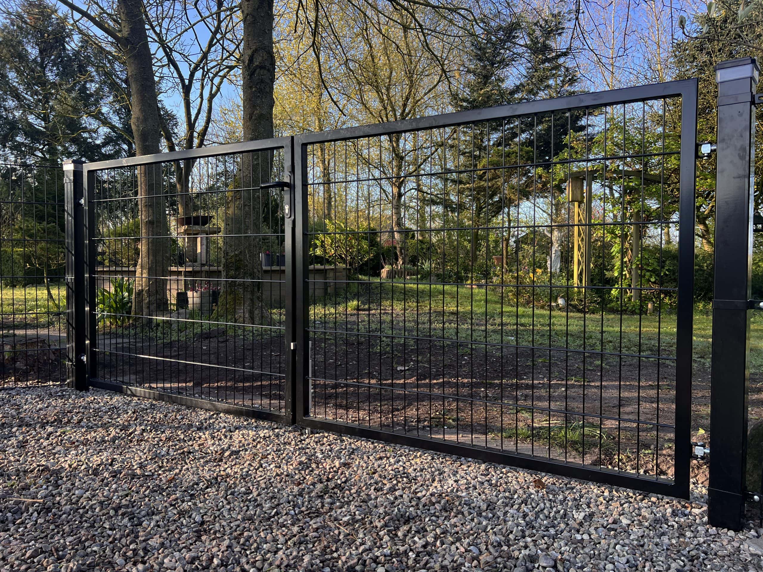 En dobbeltlåge 2D 6/5/6 mm sort RAL 9005 komplet inkl. stolper metalport med lodrette og vandrette stænger står på en grusindkørsel og åbner ud til en have med træer, græs og planter på en solskinsdag.