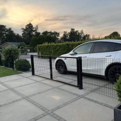 En hvid bil er parkeret bag en Metal Dobbeltlåge 3D Panel Pulverlakeret Sort RAL 9005 Komplet på en asfalteret indkørsel med grønne buske og træer i baggrunden under en overskyet aftenhimmel.