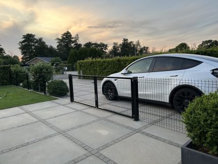 En hvid bil er parkeret bag en Metal Dobbeltlåge 3D Panel Pulverlakeret Sort RAL 9005 Komplet på en asfalteret indkørsel med grønne buske og træer i baggrunden under en overskyet aftenhimmel.