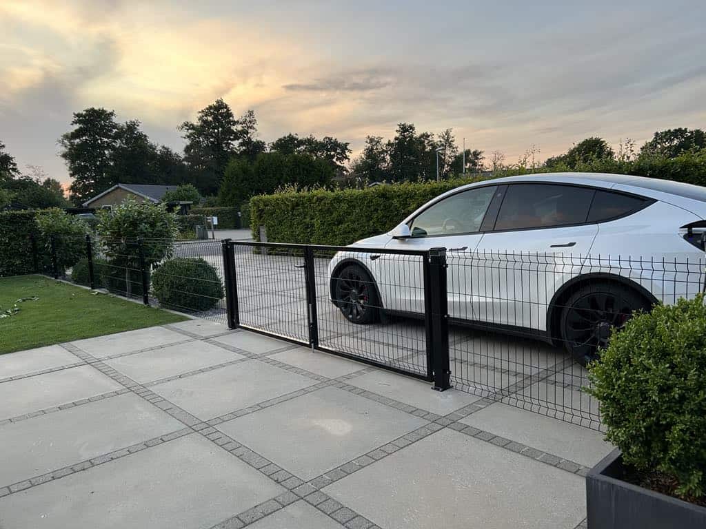 En hvid bil er parkeret bag en Metal Dobbeltlåge 3D Panel Pulverlakeret Sort RAL 9005 Komplet på en asfalteret indkørsel med grønne buske og træer i baggrunden under en overskyet aftenhimmel.