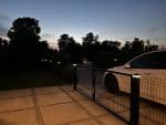 En hvid bil holder parkeret ved en Metal Dobbeltlåge 3D Panel Pulverlakeret Sort RAL 9005 Komplet i skumringen, og langs kanten lyser vejlys. Træer og hække dukker op i baggrunden under en mørkere himmel.