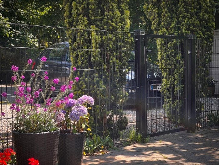 En havelåge og et hegn i sort metal med høje grønne buske bagved; en sort bil kan ses gennem hegnet. I forgrunden er der lilla og pink blomster i sorte potter på en stenterrasse.