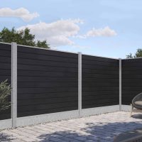 En baggård med sammenlåsende stenterrassegulve og moderne hegn. Komposit Hegn med galvaniseret bundplader, stålstolper & u-skinner – Type 2 er lavet af mørke vandrette paneler med lysegrå betonstolper og en grusplade i bunden. Der er nogle buske og træer i baggrunden, og en kurvestol er delvist synlig.