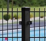Nærbillede af en sort metal hegnspæl med lodrette stænger, med en Mellemstolpe Beslag 60x40mm til Panelhegn Sort RAL 9005 (Designserie), sat i udendørs omgivelser. I baggrunden er der en swimmingpool med blåt vand og et græsareal med nogle buske. Fokus er på hegnspælen i forgrunden.
