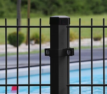 Nærbillede af en sort metal hegnspæl med lodrette stænger, med en Mellemstolpe Beslag 60x40mm til Panelhegn Sort RAL 9005 (Designserie), sat i udendørs omgivelser. I baggrunden er der en swimmingpool med blåt vand og et græsareal med nogle buske. Fokus er på hegnspælen i forgrunden.
