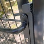 Nærbillede af håndtag og lås på en Dobbeltlåge 2D 6/5/6 mm Sort RAL 9005 Komplet Inkl. stolper, monteret på et trådhegn. Sollys filtreres gennem omgivelserne i den slørede baggrund.