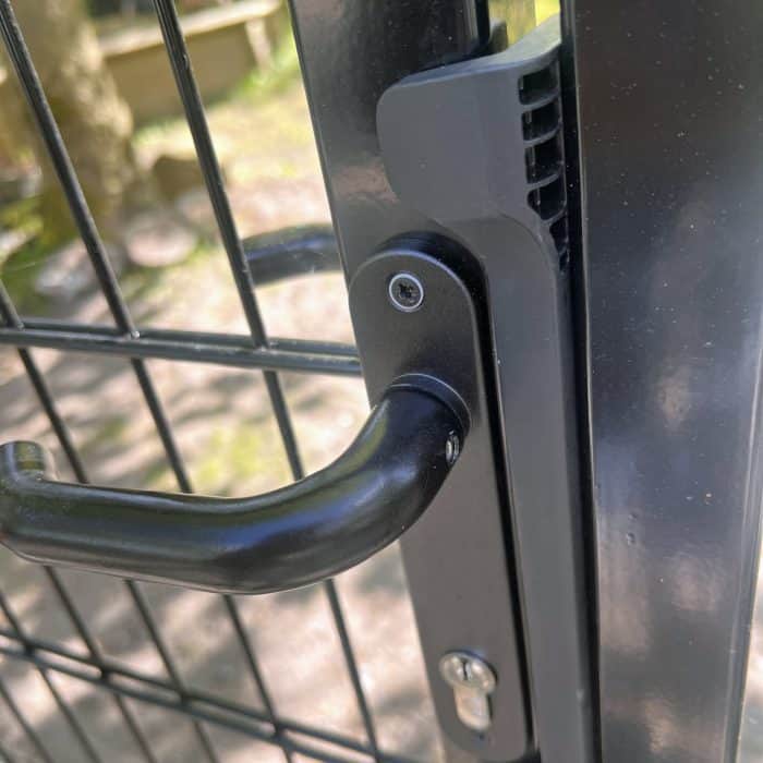 Nærbillede af håndtag og lås på en Dobbeltlåge 2D 6/5/6 mm Sort RAL 9005 Komplet Inkl. stolper, monteret på et trådhegn. Sollys filtreres gennem omgivelserne i den slørede baggrund.