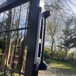 Nærbillede af en dobbeltlåge 2D 6/5/6 mm sort RAL 9005 komplet inkl. stolper portlås på et sort trådhegn med slørede træer og grønne områder i den solrige baggrund.