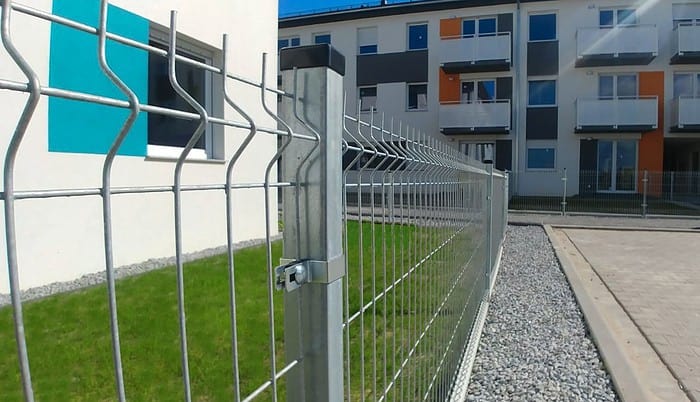 Et nærbillede af et metalhegn med en pænt trimmet grøn græsplæne på den ene side og en grussti på den anden. I baggrunden er der etageejendomme med farverige altaner, der viser den robuste Mellemstolpe Beslag 60x40mm til Panelhegn Sort RAL 9005 (Designserie), der holder det hele sammen.