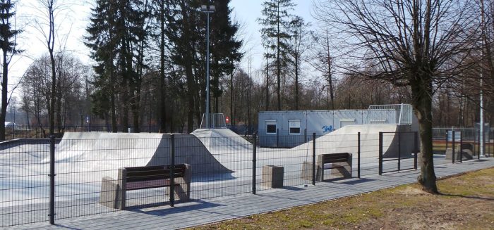 En indhegnet udendørs skatepark med betonramper og strukturer velegnet til skateboarding og BMX-cykling. Bænke placeres langs omkredsen, sikret med Mellemstolpe Beslag 60x40mm til Panelhegn Sort RAL 9005 (Designserie), mens en hvid bygning med graffiti er synlig i baggrunden. Træer omgiver området.