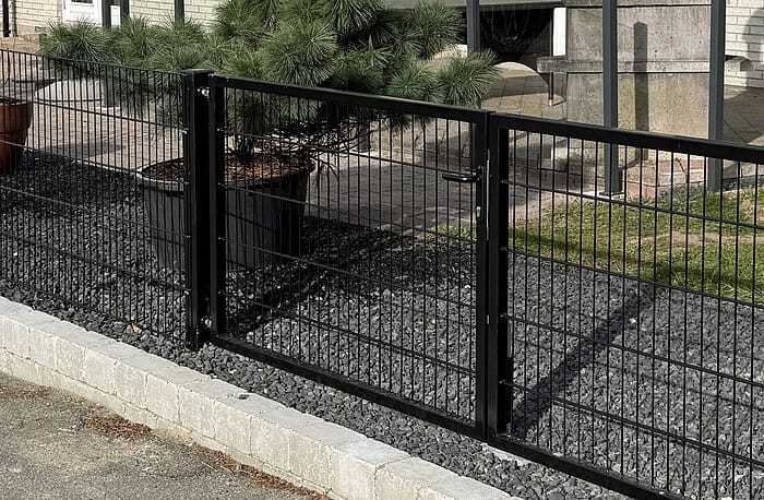 Et sort metalhegn kaldet "Dobbeltlåge 2D 6/5/6 mm Sort RAL 9005 Komplet Inkl. stolper" er monteret på en betonkant ved siden af en grussti, med et stort pottefyrtræ og græs bagved, hvilket delvist afslører en moderne bygningsindgang i baggrunden.