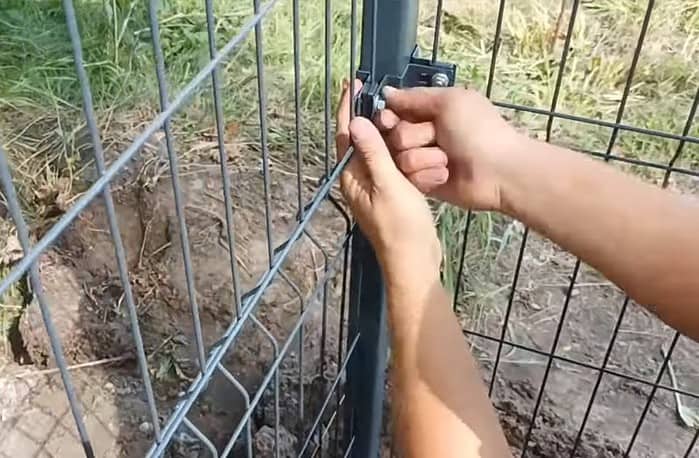 Nærbillede af hænder, der sikrer en lås på et metalhegn. Hegnet ser ud til at være i et udendørs område med græs og snavs synligt i baggrunden. Personen spænder en skrue med et værktøj, hvilket sikrer, at Hjørnestolpe Beslag 60x40mm til Panelhegn Sort RAL 9005 (Designserie) sidder godt fast.