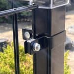 Nærbillede af en sort metalhegnspæl med en bolt og Hjørnestolpe Beslag 60x40mm til Panelhegn Sort RAL 9005 (Designserie), der fastgør to paneler, mod grønt område og en sløret, solbeskinnet baggrund.