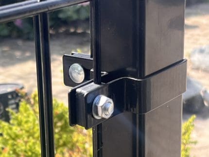 Nærbillede af en sort metalhegnspæl med en bolt og Hjørnestolpe Beslag 60x40mm til Panelhegn Sort RAL 9005 (Designserie), der fastgør to paneler, mod grønt område og en sløret, solbeskinnet baggrund.