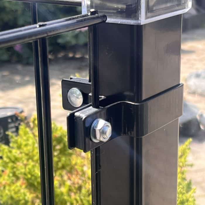 Nærbillede af en sort metalhegnspæl med en bolt og Hjørnestolpe Beslag 60x40mm til Panelhegn Sort RAL 9005 (Designserie), der fastgør to paneler, mod grønt område og en sløret, solbeskinnet baggrund.