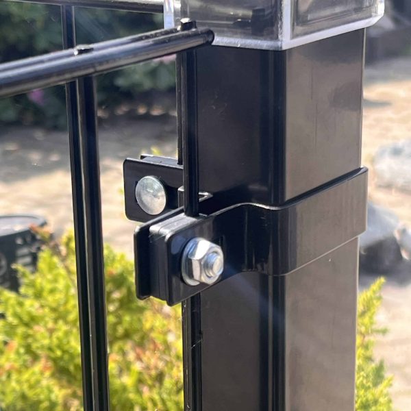 Nærbillede af en sort metalhegnspæl med en bolt og Hjørnestolpe Beslag 60x40mm til Panelhegn Sort RAL 9005 (Designserie), der fastgør to paneler, mod grønt område og en sløret, solbeskinnet baggrund.