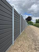 En lang, høj, mørkegrå Komposit Hegn med galvaniseret bundplader lavet af vandrette paneler er forankret af lodrette stålstolper & u-skinner. Hegnet grænser op til en jordvej med pletter af græs. I baggrunden er der træer, en hæk og en overskyet himmel.