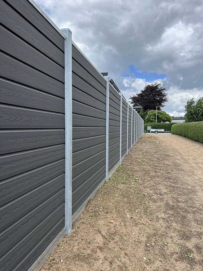 En lang, høj, mørkegrå Komposit Hegn med galvaniseret bundplader lavet af vandrette paneler er forankret af lodrette stålstolper & u-skinner. Hegnet grænser op til en jordvej med pletter af græs. I baggrunden er der træer, en hæk og en overskyet himmel.
