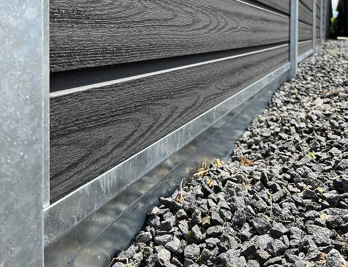 Nærbillede af bunden af en mørk træbeklædt væg med metalbeklædning i bunden, støder op til en jord dækket med mørkegrå grus. Træets tekstur og ruheden af gruset er tydeligt synlige, hvilket fremhæver kontrasten mellem materialer i denne Komposit Hegn med galvaniseret bundplader, stålstolper & u-skinner – Type 2.