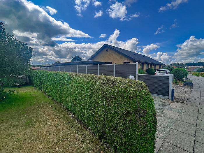 En velholdt hæk og en Komposit Hegn med galvaniseret bundplader, stålstolper & u-skinner – Type 2 langs en forstadssti, der fører til et moderne murstenshus med skrånende tag. Himlen er delvist overskyet med blå pletter, og en bil er parkeret i indkørslen. Scenen er fredelig og velordnet.