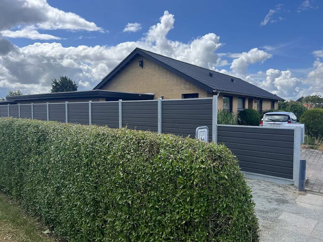 Et moderne etplanshus med murstensfacade og sort tegltag er delvist sløret af en høj, grøn hæk og en Komposit Hegn med galvaniseret bundplader, stålstolper & u-skinner – Type 2. En indkørsel fører til en garage med en hvid bil parkeret bag porten. Himlen er delvist overskyet.