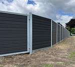 Et langt, moderne gråt hegn lavet af vandrette paneler med metalstolper løber langs en jordsti ved siden af græsset under en overskyet himmel. Denne Komposit Hegn med galvaniseret bundplader, stålstolper & u-skinner – Type 2 står fast, da træer og buske kan ses i det fjerne.