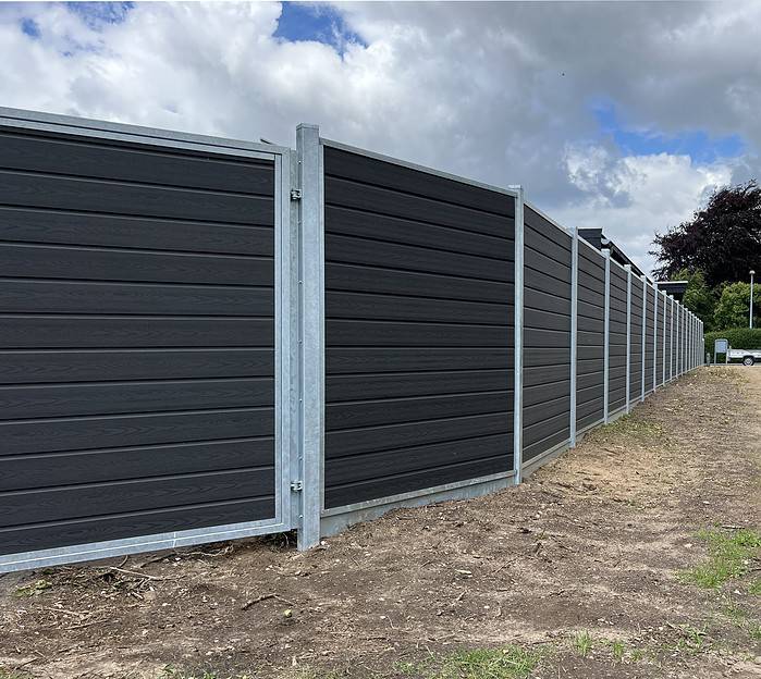 Et langt, moderne gråt hegn lavet af vandrette paneler med metalstolper løber langs en jordsti ved siden af græsset under en overskyet himmel. Denne Komposit Hegn med galvaniseret bundplader, stålstolper & u-skinner – Type 2 står fast, da træer og buske kan ses i det fjerne.