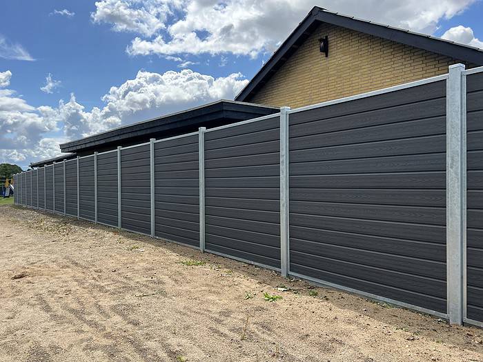 Et højt, mørkegrå vinylhegn med metalpæle strækker sig langs omkredsen af en ejendom og giver privatlivets fred fra naboområderne. Denne Komposit Hegn med galvaniseret bundplader, stålstolper & u-skinner – Type 2 står ved siden af en moderne murstensbygning med skrånende tag, og himlen ovenover er delvist overskyet.