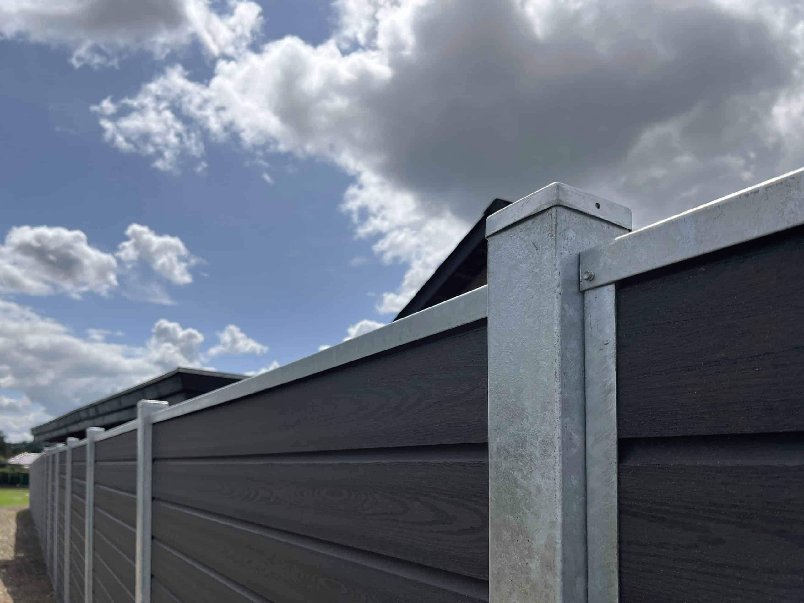 Et nærbillede af en høj, mørkfarvet Komposit Hegn med galvaniseret bundplader, stålstolper & u-skinner – Type 2 opbygget af vandrette paneler og metalstolper, under en delvist overskyet himmel. Solen oplyser dele af hegnet og fremhæver dets tekstur.
