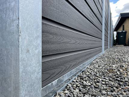 Nærbillede af siden af et moderne hus med mørk vandret sidebeklædning med metalkant i bunden. Grunden nedenunder er dækket af småsten, ved siden af et afsnit med Komposit Hegn med galvaniseret bundplader, stålstolper & u-skinner – Type 2. En fjern del af huset og en sort skraldespand er synlige nede ad stien. Himlen er delvist overskyet.