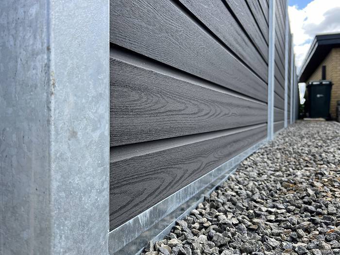 Nærbillede af siden af et moderne hus med mørk vandret sidebeklædning med metalkant i bunden. Grunden nedenunder er dækket af småsten, ved siden af et afsnit med Komposit Hegn med galvaniseret bundplader, stålstolper & u-skinner – Type 2. En fjern del af huset og en sort skraldespand er synlige nede ad stien. Himlen er delvist overskyet.