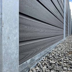 Nærbillede af siden af et moderne hus med mørk vandret sidebeklædning med metalkant i bunden. Grunden nedenunder er dækket af småsten, ved siden af et afsnit med Komposit Hegn med galvaniseret bundplader, stålstolper & u-skinner – Type 2. En fjern del af huset og en sort skraldespand er synlige nede ad stien. Himlen er delvist overskyet.