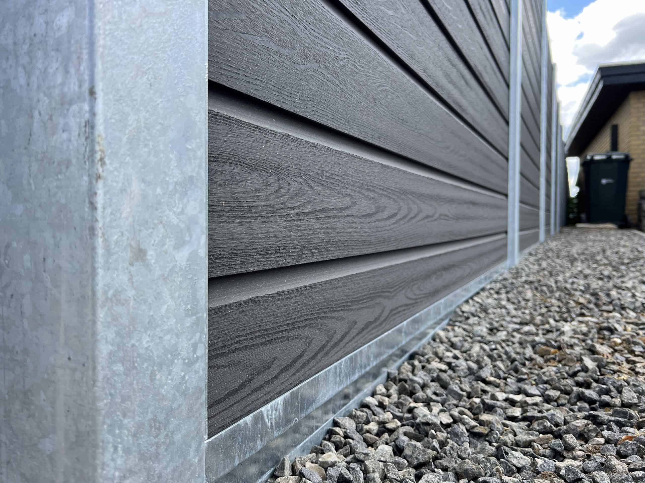 Nærbillede af siden af et moderne hus med mørk vandret sidebeklædning med metalkant i bunden. Grunden nedenunder er dækket af småsten, ved siden af et afsnit med Komposit Hegn med galvaniseret bundplader, stålstolper & u-skinner – Type 2. En fjern del af huset og en sort skraldespand er synlige nede ad stien. Himlen er delvist overskyet.