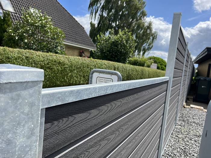 En nyinstalleret, moderne Komposit Hegn med galvaniseret bundplader, stålstolper & u-skinner – Type 2 står ved siden af en pænt trimmet hæk i et forstadskvarter. Himlen er delvist overskyet med blå pletter, og et hus med skrånende tag og mere grønt er synligt i baggrunden.