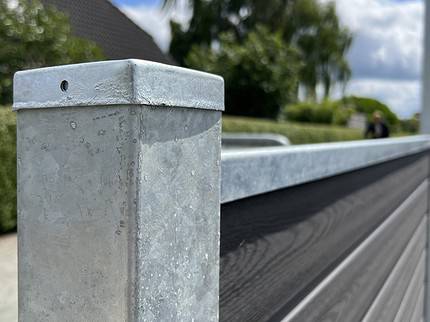 Et nærbillede af en metallisk hegnspæl med en afdækket top og en vandret bjælke, der strækker sig ud fra den, viser teksturen og strukturen af Komposit Hegn med galvaniseret bundplader, stålstolper & u-skinner – Type 2. Baggrunden har sløret grønt, et hus , og en blå himmel med spredte skyer.