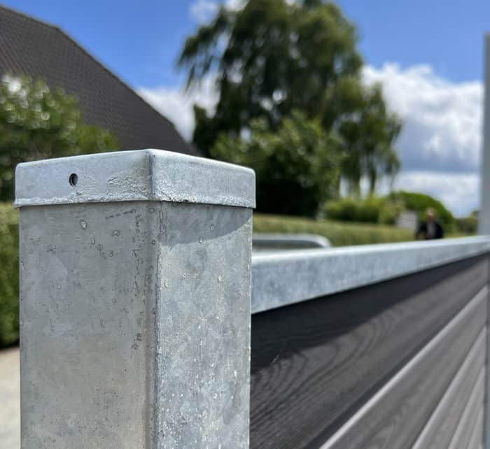Et nærbillede af en metallisk hegnspæl med en afdækket top og en vandret bjælke, der strækker sig ud fra den, viser teksturen og strukturen af Komposit Hegn med galvaniseret bundplader, stålstolper & u-skinner – Type 2. Baggrunden har sløret grønt, et hus , og en blå himmel med spredte skyer.