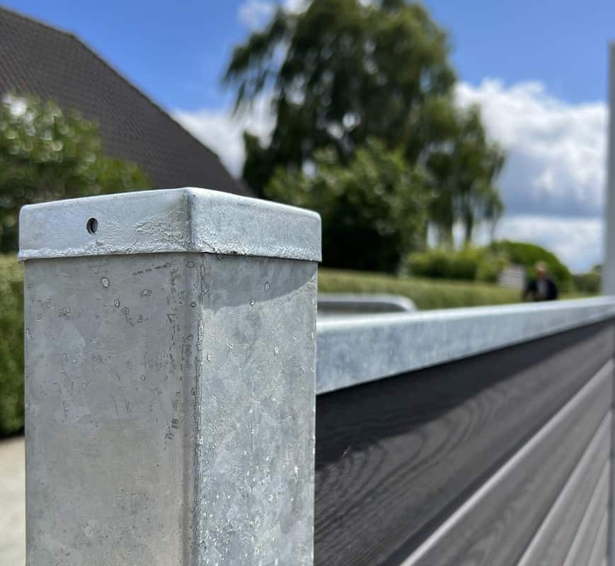 Et nærbillede af en metallisk hegnspæl med en afdækket top og en vandret bjælke, der strækker sig ud fra den, viser teksturen og strukturen af Komposit Hegn med galvaniseret bundplader, stålstolper & u-skinner – Type 2. Baggrunden har sløret grønt, et hus , og en blå himmel med spredte skyer.