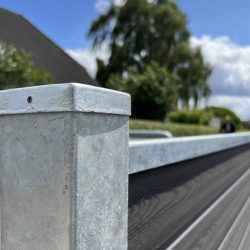 Et nærbillede af en metallisk hegnspæl med en afdækket top og en vandret bjælke, der strækker sig ud fra den, viser teksturen og strukturen af Komposit Hegn med galvaniseret bundplader, stålstolper & u-skinner – Type 2. Baggrunden har sløret grønt, et hus , og en blå himmel med spredte skyer.