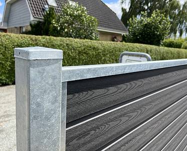 Nærbillede af en Komposit Hegn med galvaniseret bundplader, stålstolper & u-skinner – Type 2, med vandrette trælignende paneler indrammet af en galvaniseret metalstolpe og topskinne. Hegnet er placeret udendørs med en pænt klippet hæk og et hus med tegltag i baggrunden under en solrig himmel.